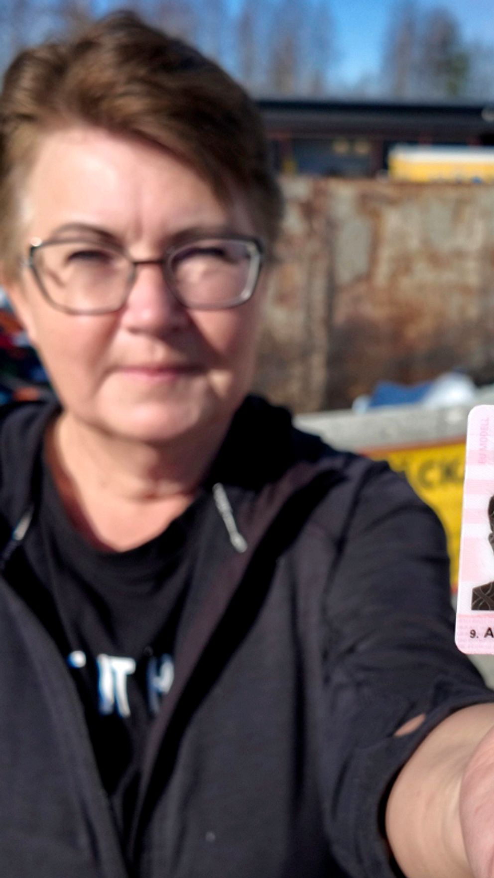 Lena Bitén håller upp ett körkort mot kameran. Bakgrund med containrar och skrot.