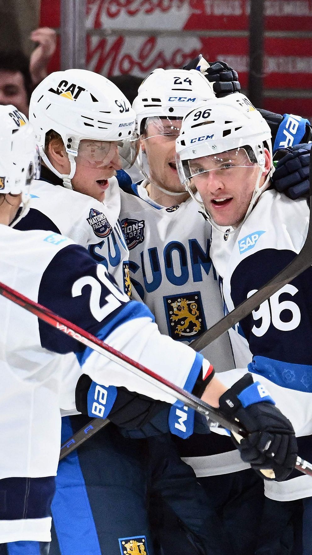 Fem ishockeyspelare från Finland firar ett mål på isen med sina klubbor uppsträckta, iklädda vit-blåa matchtröjor med texten ”SUOMI”. I bakgrunden syns en ishockeyrinks skyltar.