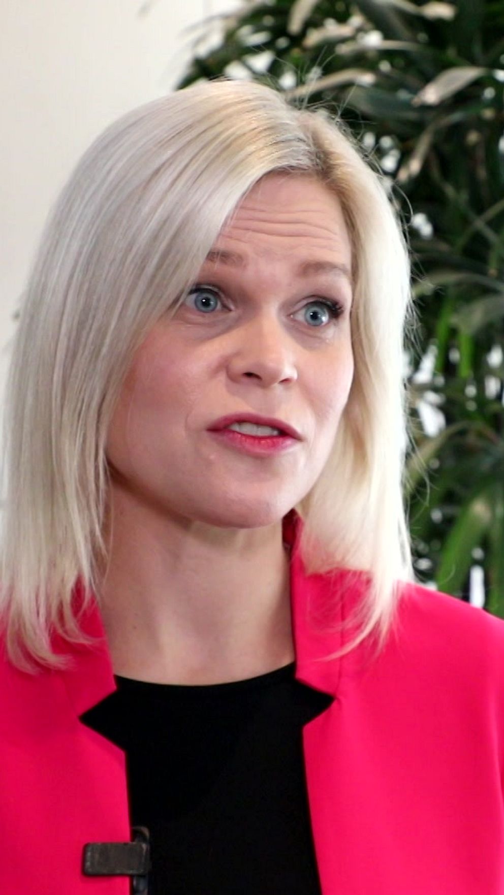 En person står framför en växt i en ljus inomhusmiljö, klädd i en röd jacka över en svart topp. Bakgrunden har flera stora gröna växter och en ljus vägg som bidrar till en professionell atmosfär.