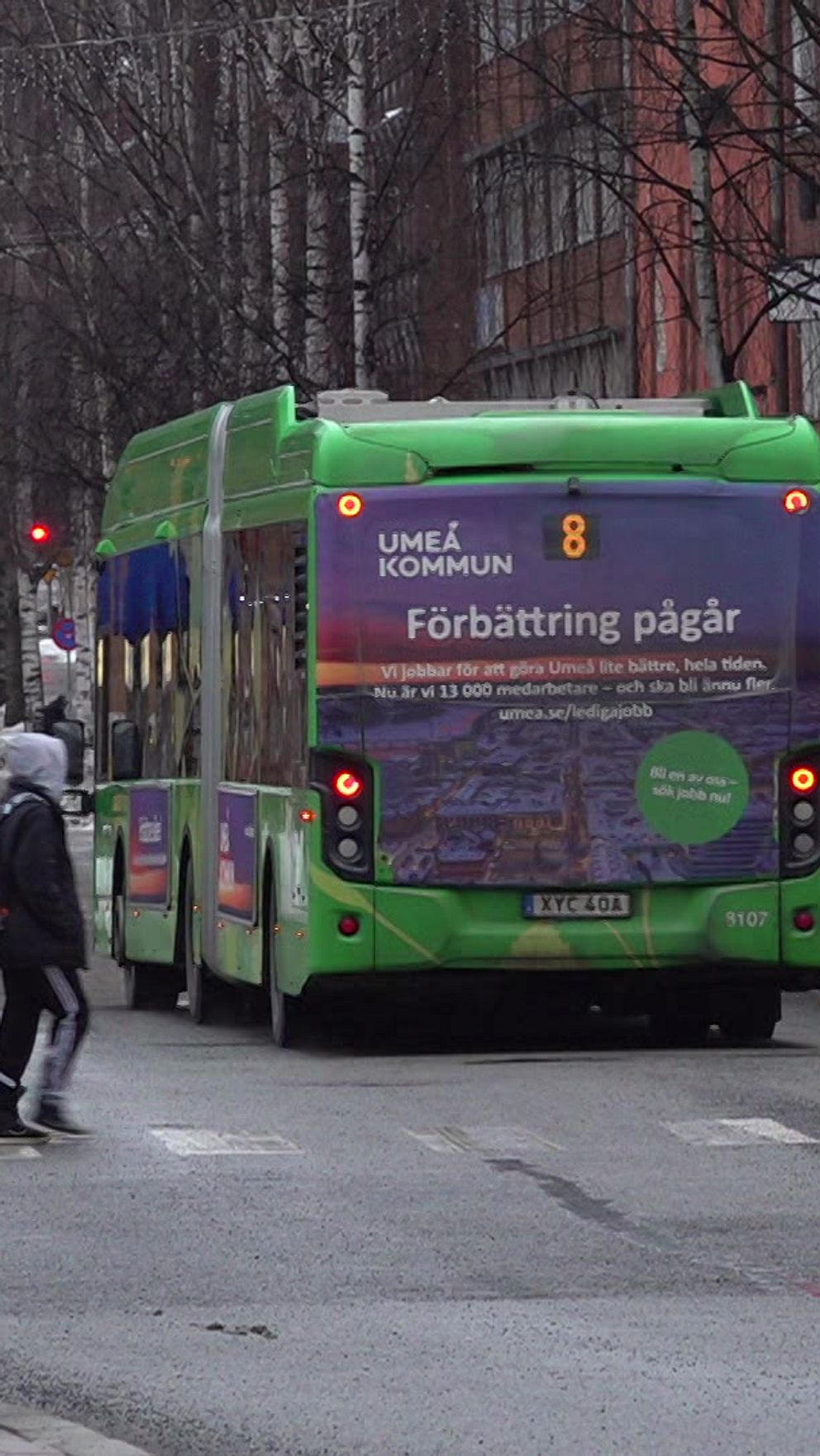 En grön och lila stadsbuss som kör förbi en övergångsställe medan flera personer går över gatan. I bakgrunden syns byggnader och en snöig gata, vilket signalerar vintermånaderna. Bussens bakdel har texten ”Förbättring pågår” vilket antyder att det pågår arbeten eller förändringar i området.