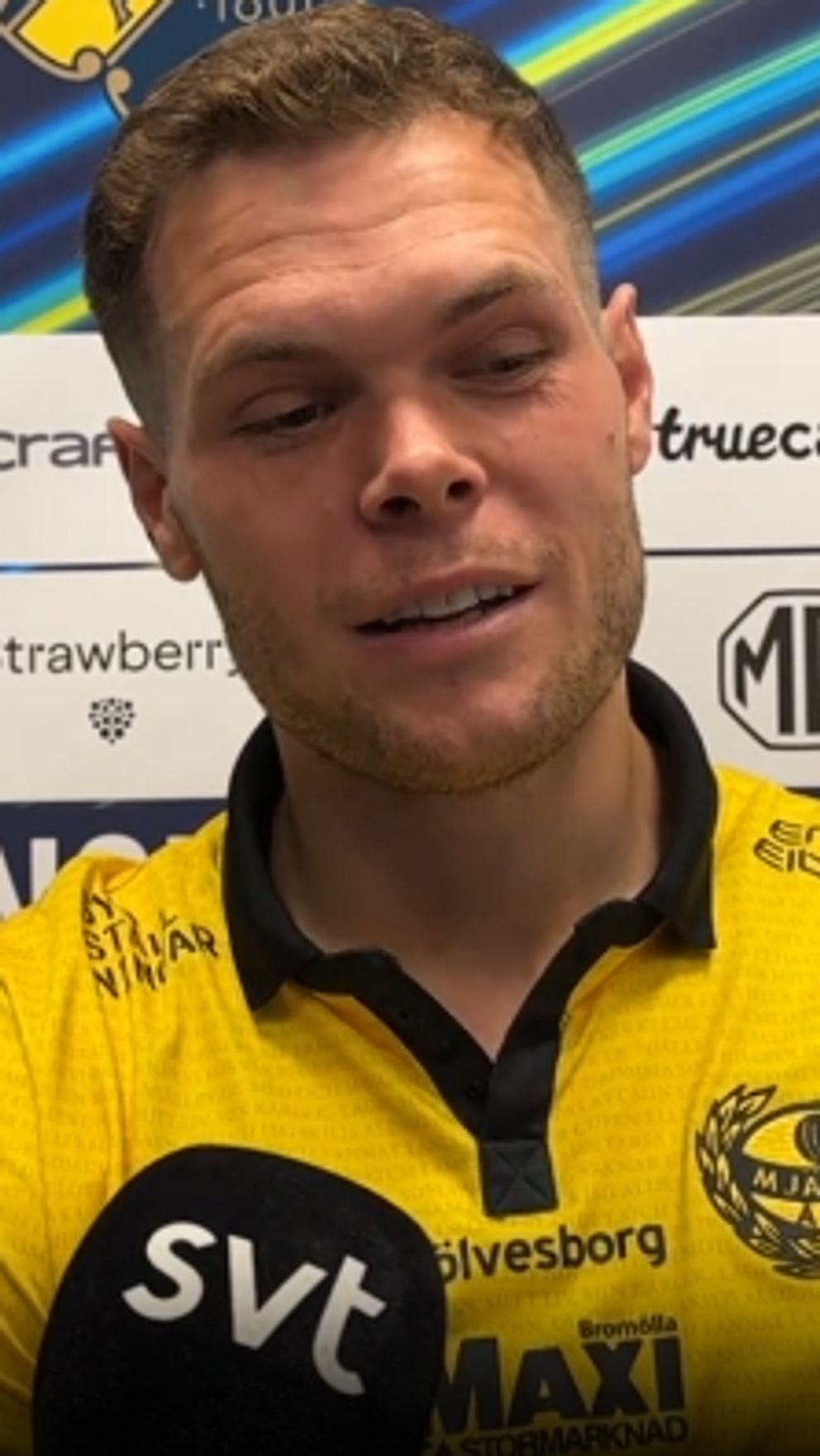 Person i gul tröja intervjuas av SVT med sponsorvägg i bakgrunden.