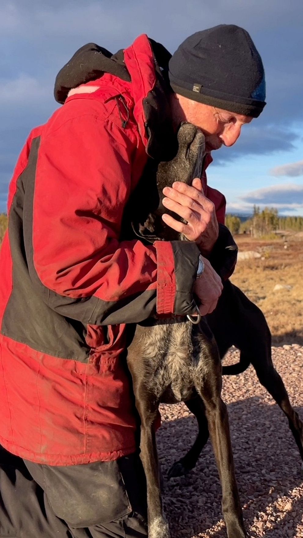 Man i röd jacka kramar en hund på grusväg under blå himmel.
