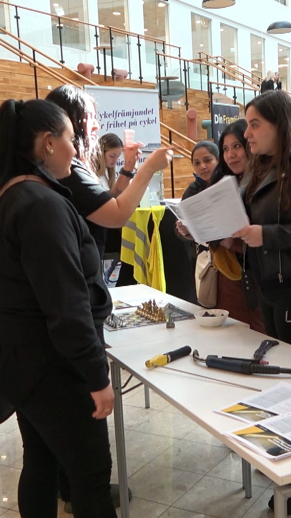 En grupp personer samlas vid en monter på en mässa eller ett event. De diskuterar information och deltar i en interaktion med representanter som presenterar material och broschyrer. I bakgrunden syns en stor skärm som visar innehåll relaterat till evenemanget.