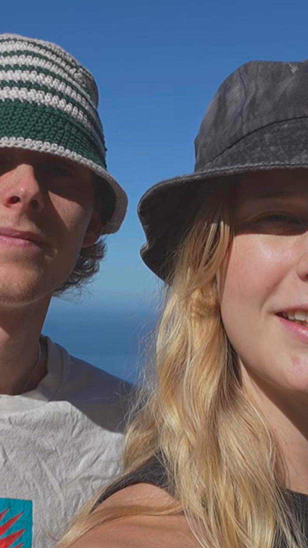 Två personer poserar framför en panoramautsikt över havet, båda med hattar och leenden. Den ena bär en vit t-shirt med färgglad design, medan den andra har på sig en svart topp. I bakgrunden syns en klar blå himmel och klippiga kustlinjer.