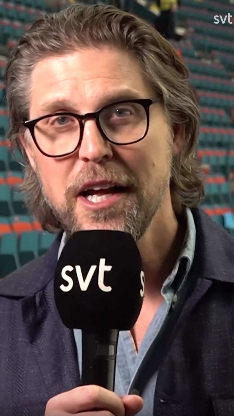Person håller SVT-mikrofon, bakgrund med tomma läktarstolar.