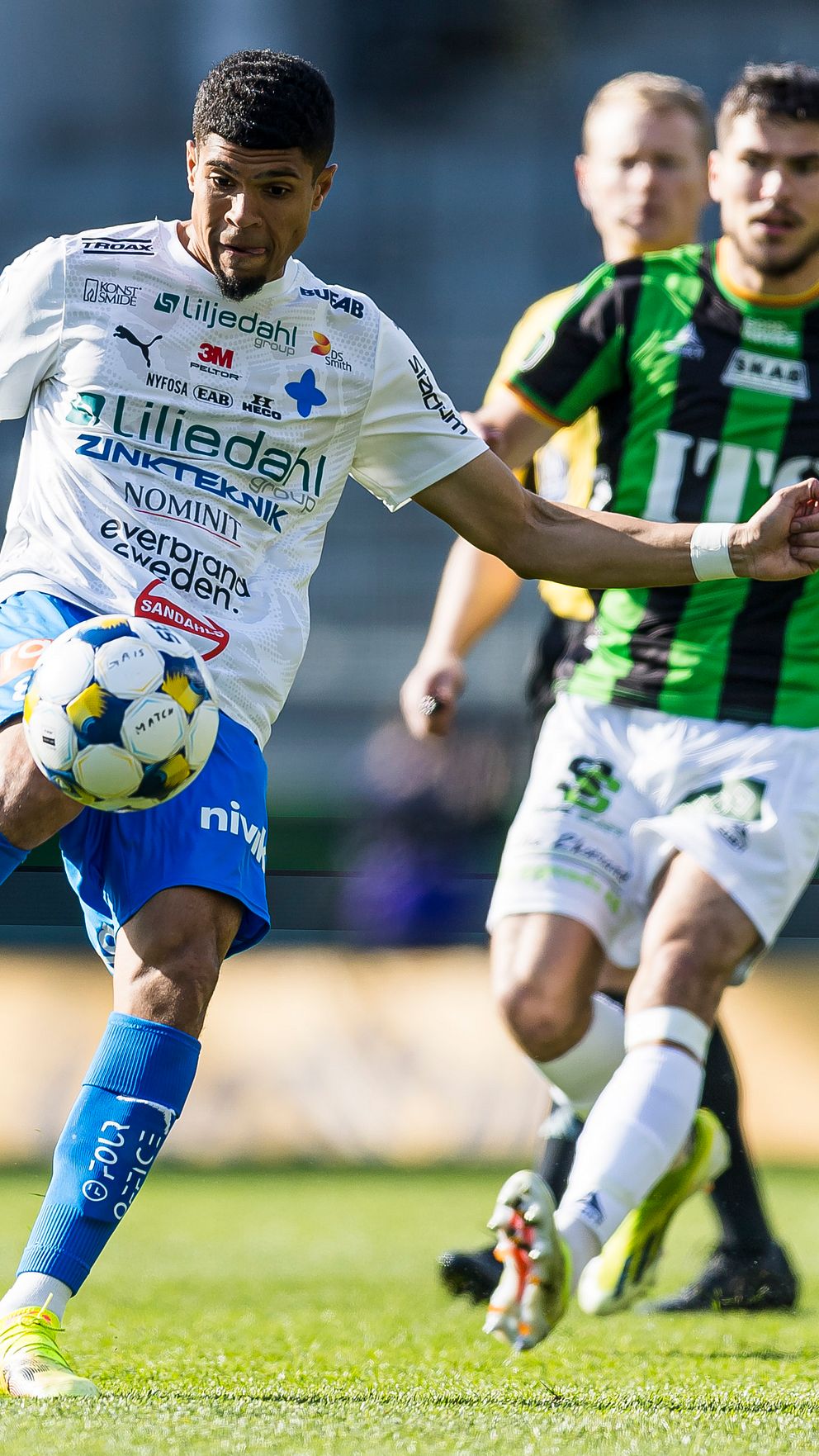 Fotbollsspelare i vit och blå dräkt sparkar bollen, motståndare i grönsvart förföljer.