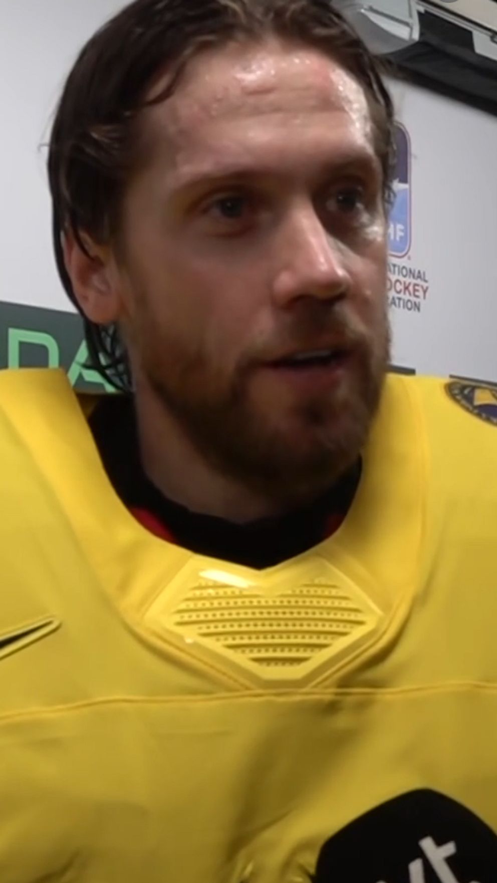 Ishockeyspelare i gul landslagströja intervjuas framför sponsortavla.