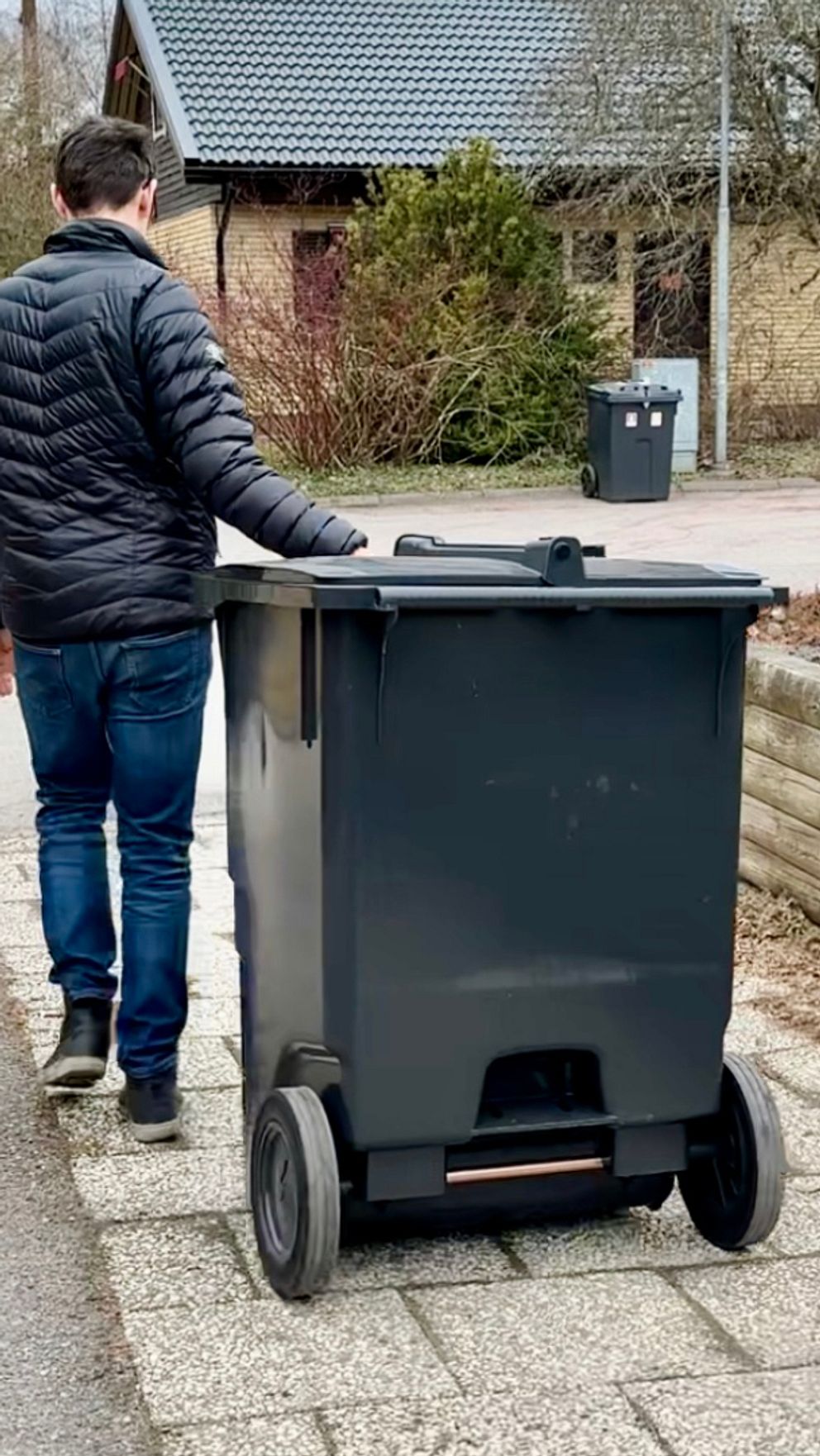Person i svart jacka drar ett soptunna längs en trottoar i ett bostadsområde.