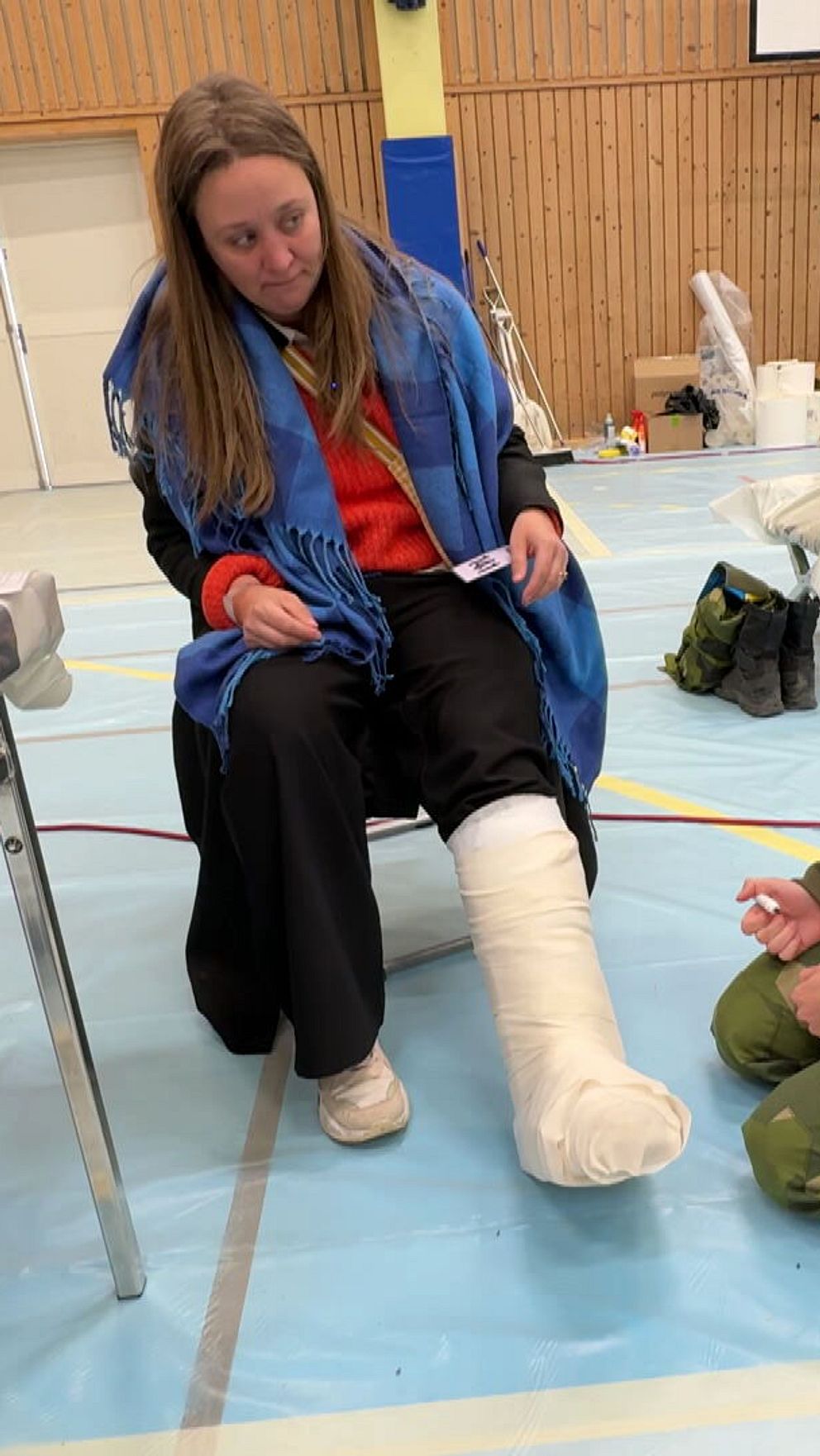 Kvinna med gipsat ben sitter, sjukvårdare i militäruniform knäar bredvid henne i idrottshall.
