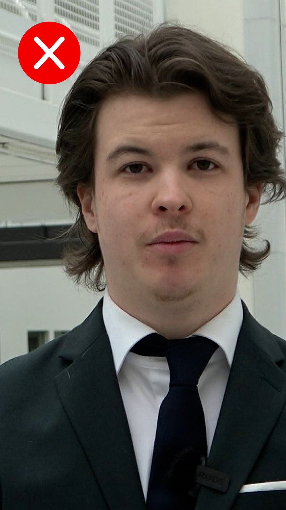 Man i kostym och slips med ett inomhusmiljö i bakgrunden. I hörnet är det ett rött kryss.