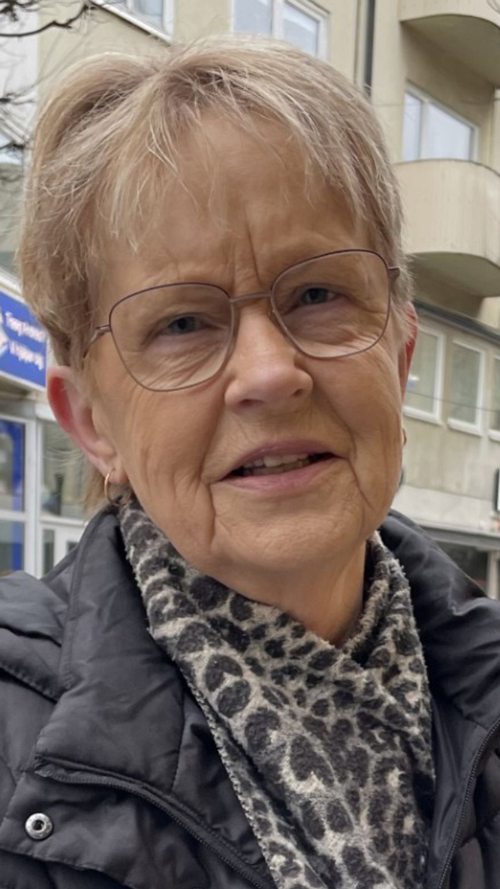 En kvinna står på en stadsgata med affärer och restauranger i bakgrunden. Träd och planteringar pryder gatan där även förbipasserande går.