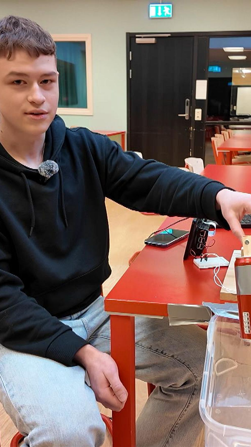 En person sitter vid ett rött bord i ett klassrum och häller något från en röd kartong ner i en genomskinlig plastbehållare. Runt omkring finns fler tomma bord och stolar.