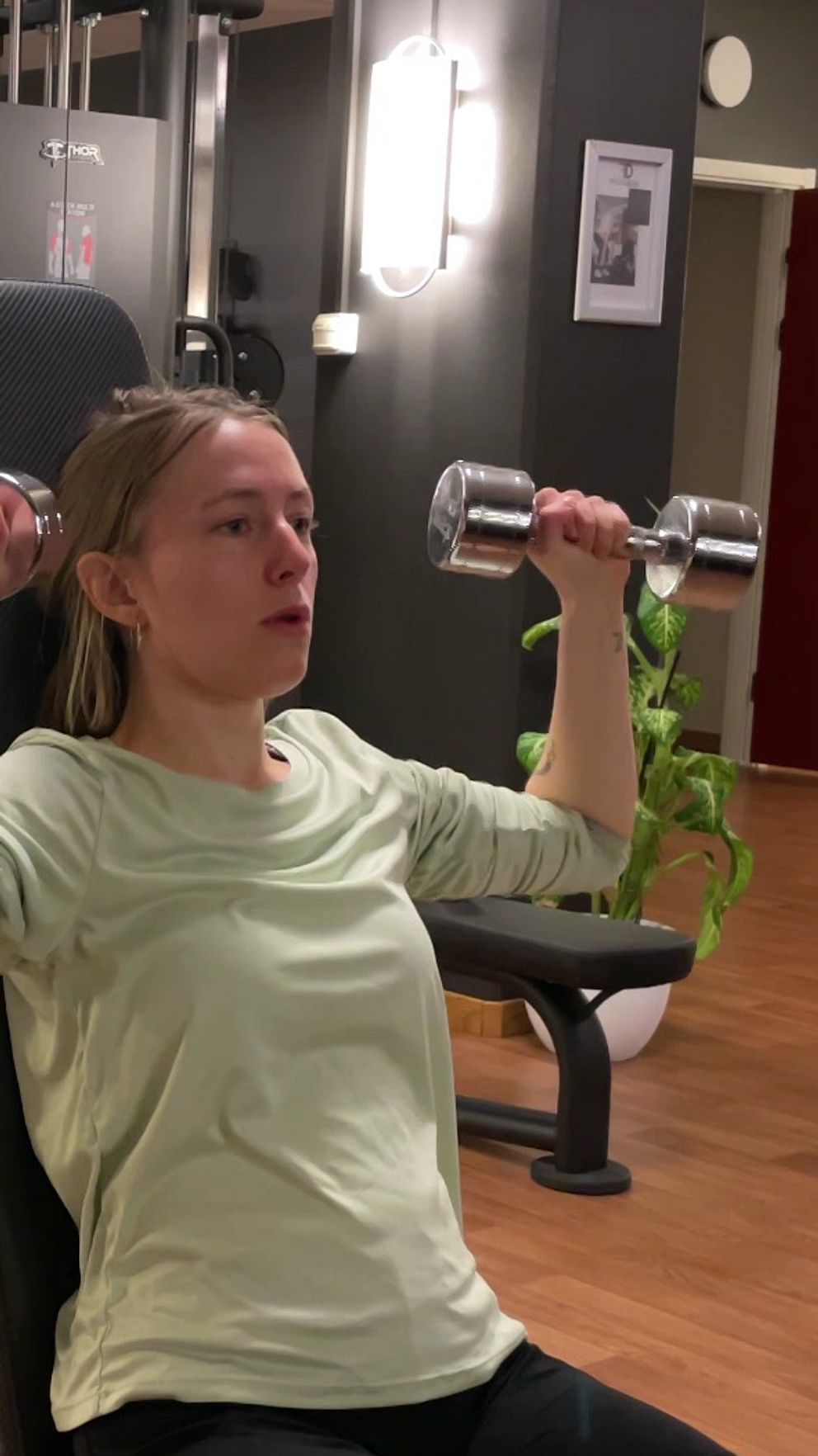 En person i en grön tröja tränar med hantlar i en gymmiljö. I bakgrunden syns träningsmaskiner, en öppen dörr och en krukväxt längs en väg med trägolv.