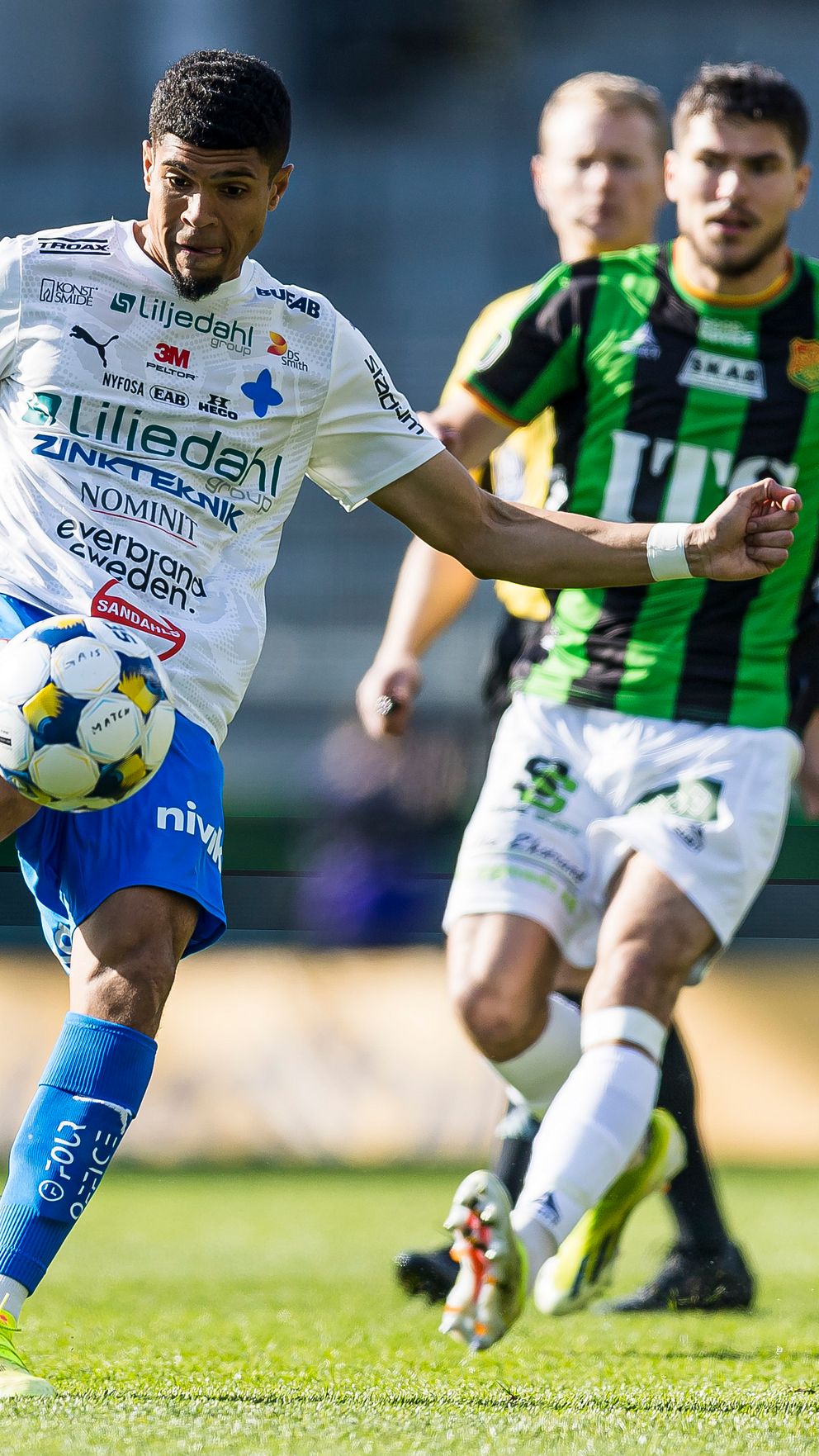 Fotbollsspelare i vit och blå dräkt sparkar bollen, motståndare i grönsvart förföljer.
