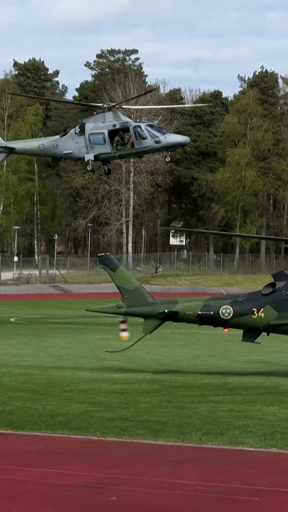 Två militärhelikoptrar landar på en idrottsplan omgiven av skog och löparbana.