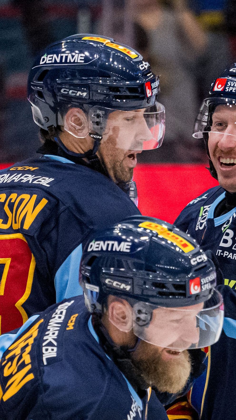 Tre ishockeyspelare i matchtröjor står på isen, samlade i en situation som verkar vara efter ett spel. Den första spelaren bär nummer 48, den andra nummer 82 och den tredje nummer 41. De har på sig hjälmar och håller i sina klubbor. Bakom dem syns en diffus bakgrund av en ishockeyarena.
