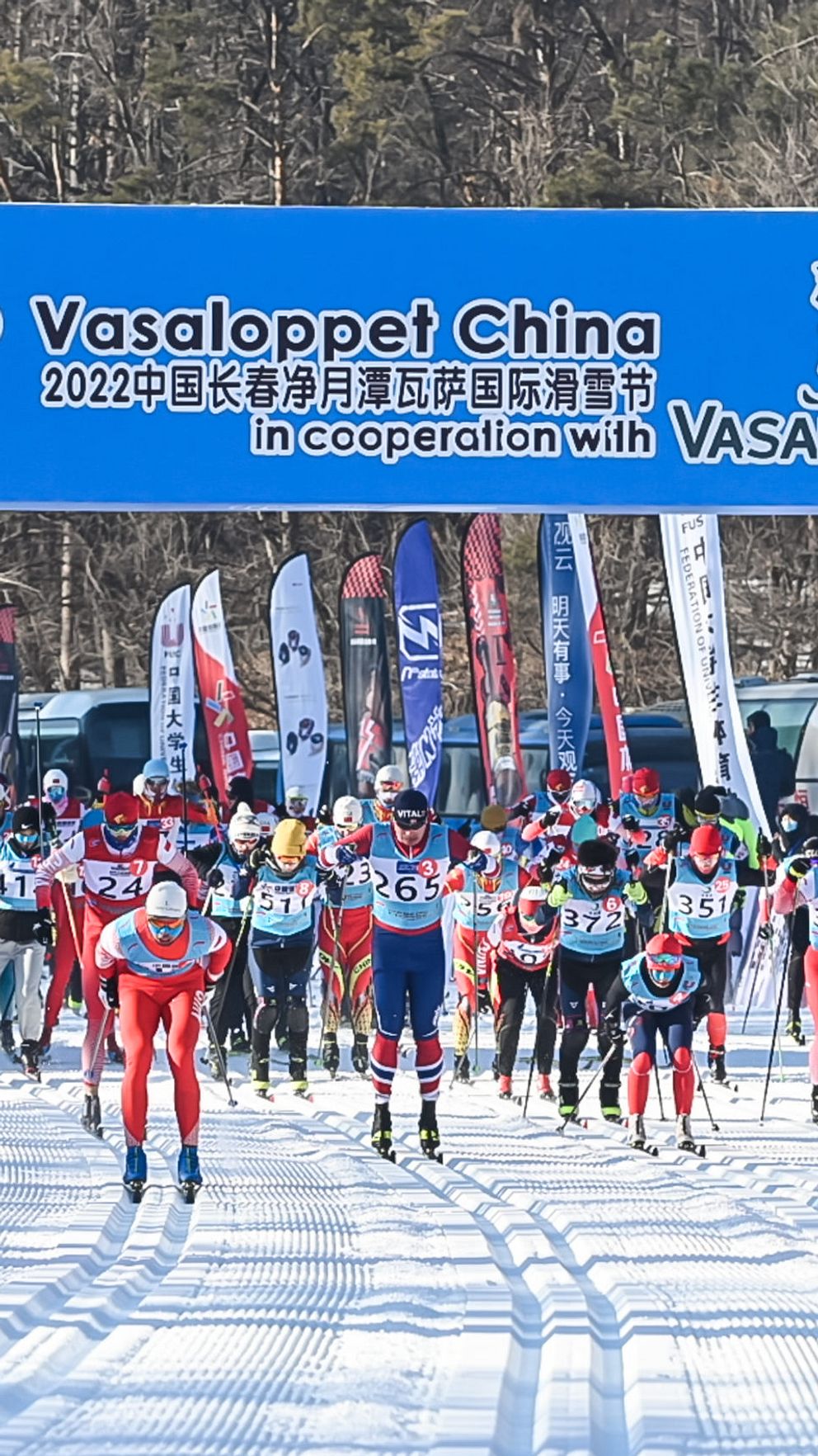 Flera skidlöpare i färgglada dräkter börjar starta i Vasaloppet China 2022, med en stor banner i bakgrunden som visar evenemangets namn och samarbetspartners. Omkring dem finns åskådare och flaggor som markerar starten för loppet.