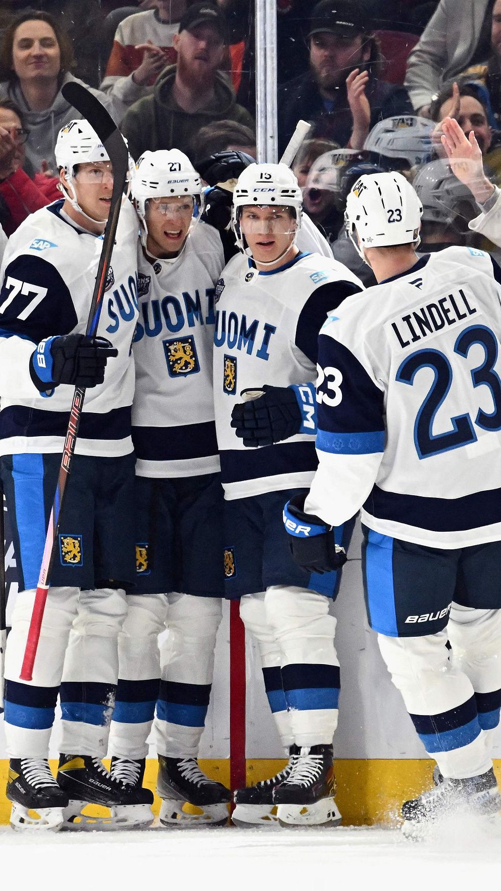 Fyra ishockeyspelare från Finland firar ett mål framför en glad publik under en match. Spelarna har på sig sina landslagsdräkter och står tillsammans nära rinken medan fansen klappar och fotar i bakgrunden.