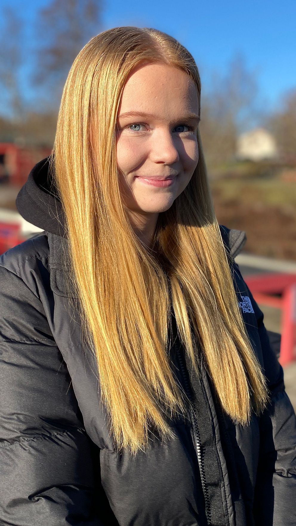 En person med långt blont hår står ute i en park eller offentlig plats, klädd i en svart, vadderad jacka. Bakgrunden visar en blå himmel och kala träd i dagsljus. En röd konstruktion, kanske en bro eller räcke, syns i bakgrunden.