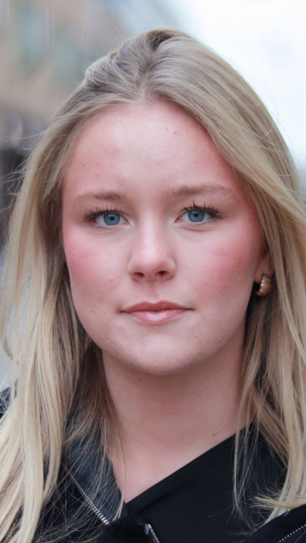 En kvinna med långt blont hår står på en gata med människor och affärer i bakgrunden. Gatan är suddig och ger ett intryck av en livlig stadsatmosfär.