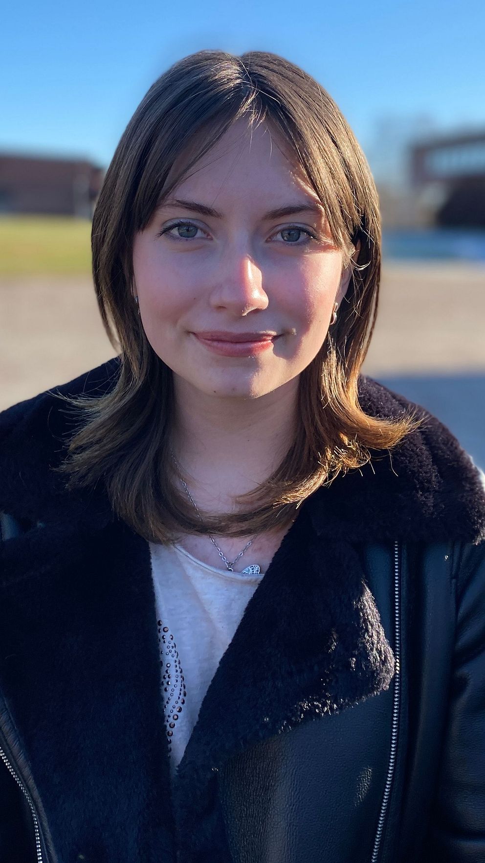 En kvinna i en svart jacka står på en solig trottoar med en byggnad och gräsmatta i bakgrunden.
