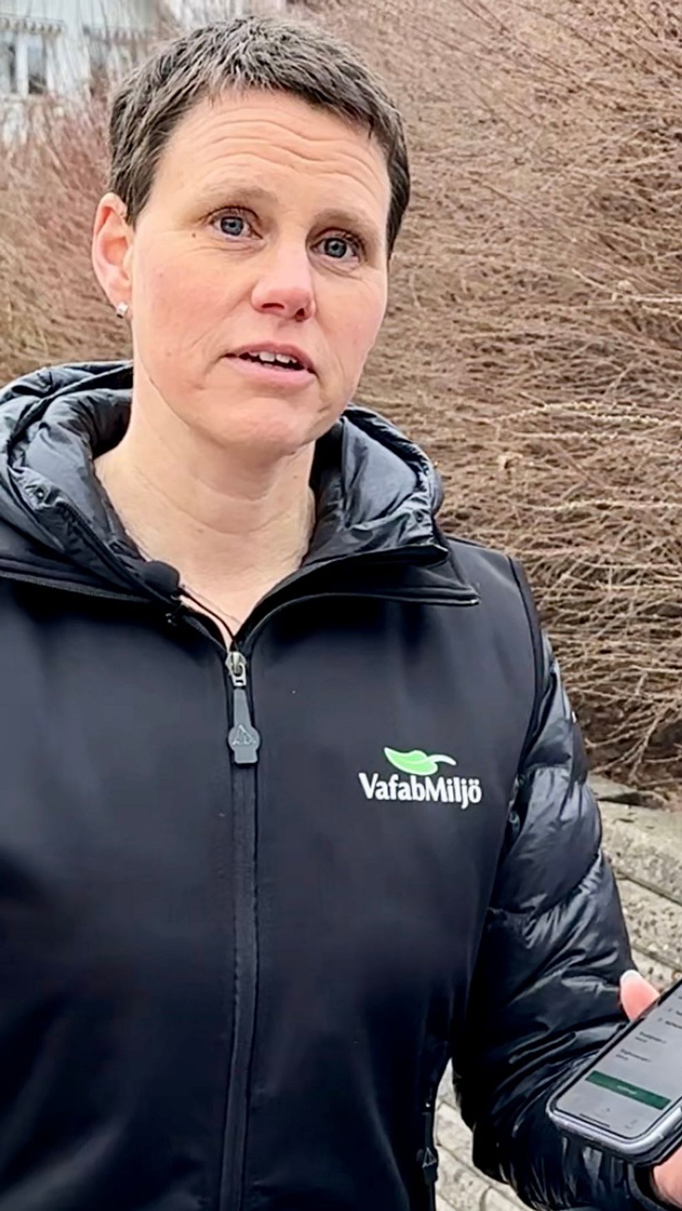 Person i svart jacka håller telefon vid buskage. Papperskorg syns i bakgrunden.