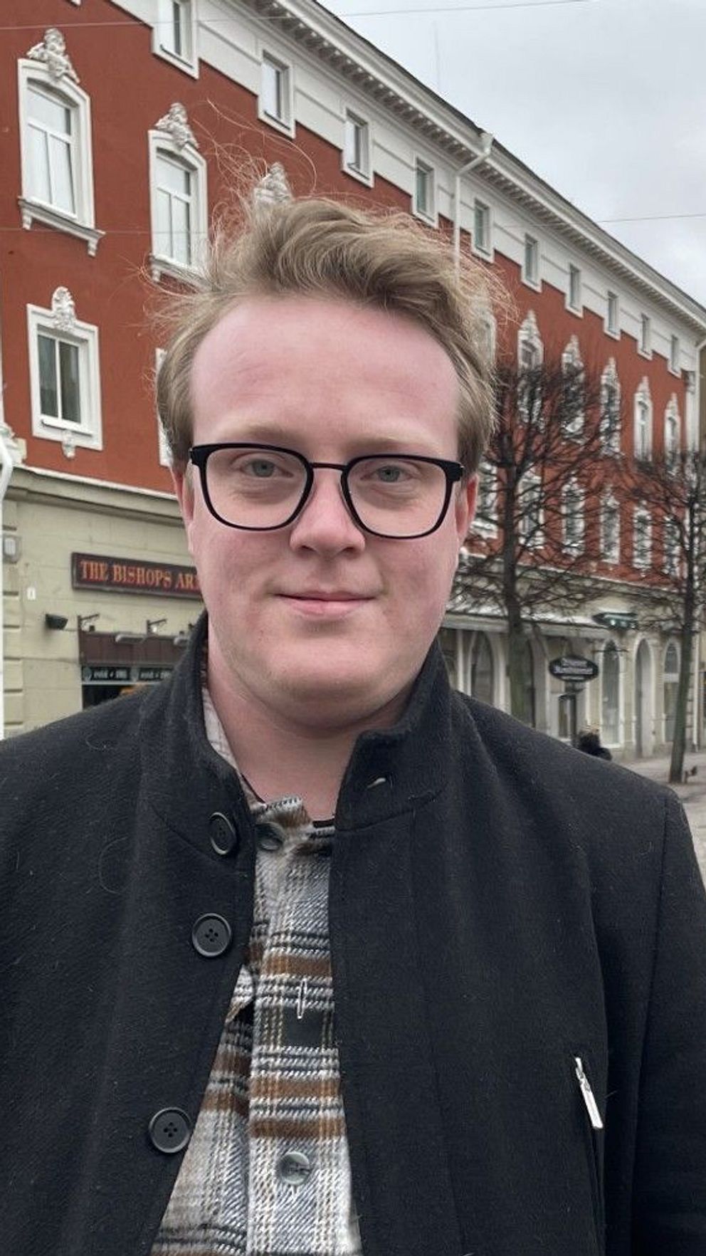 En man står på en kullerstensgata. Bakom mannen syns byggnader och människor som går och cyklar.