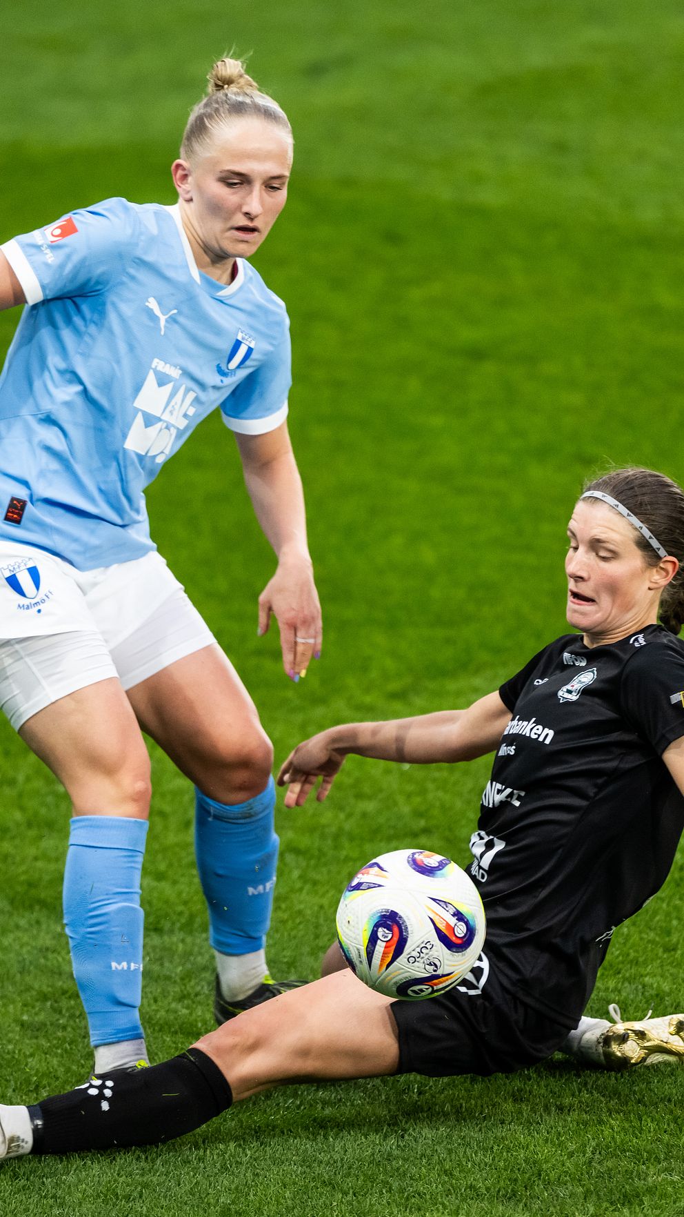 En spelare i svart tacklar en spelare i ljusblått under en fotbollsmatch på gräsplan.