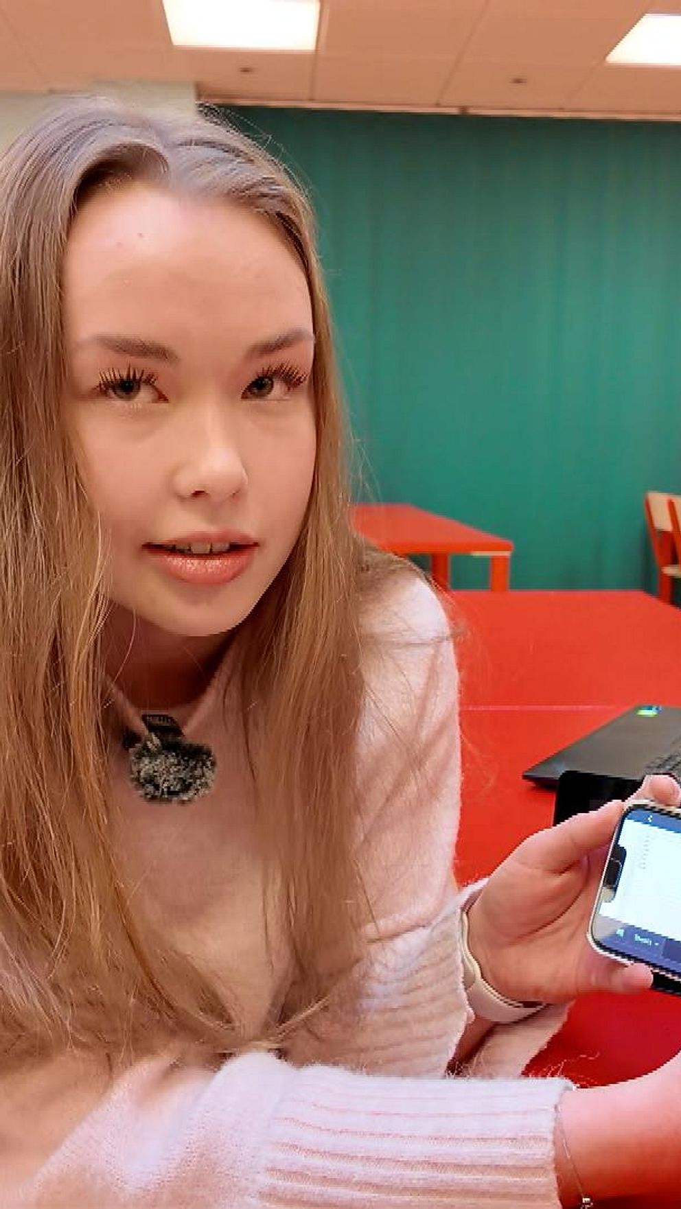 En person i en tom klassrumsmiljö sitter vid ett rött bord och håller upp en smartphone som visar en vit skärm. I bakgrunden finns fler röda bord och stolar samt en bärbar dator på bordet.