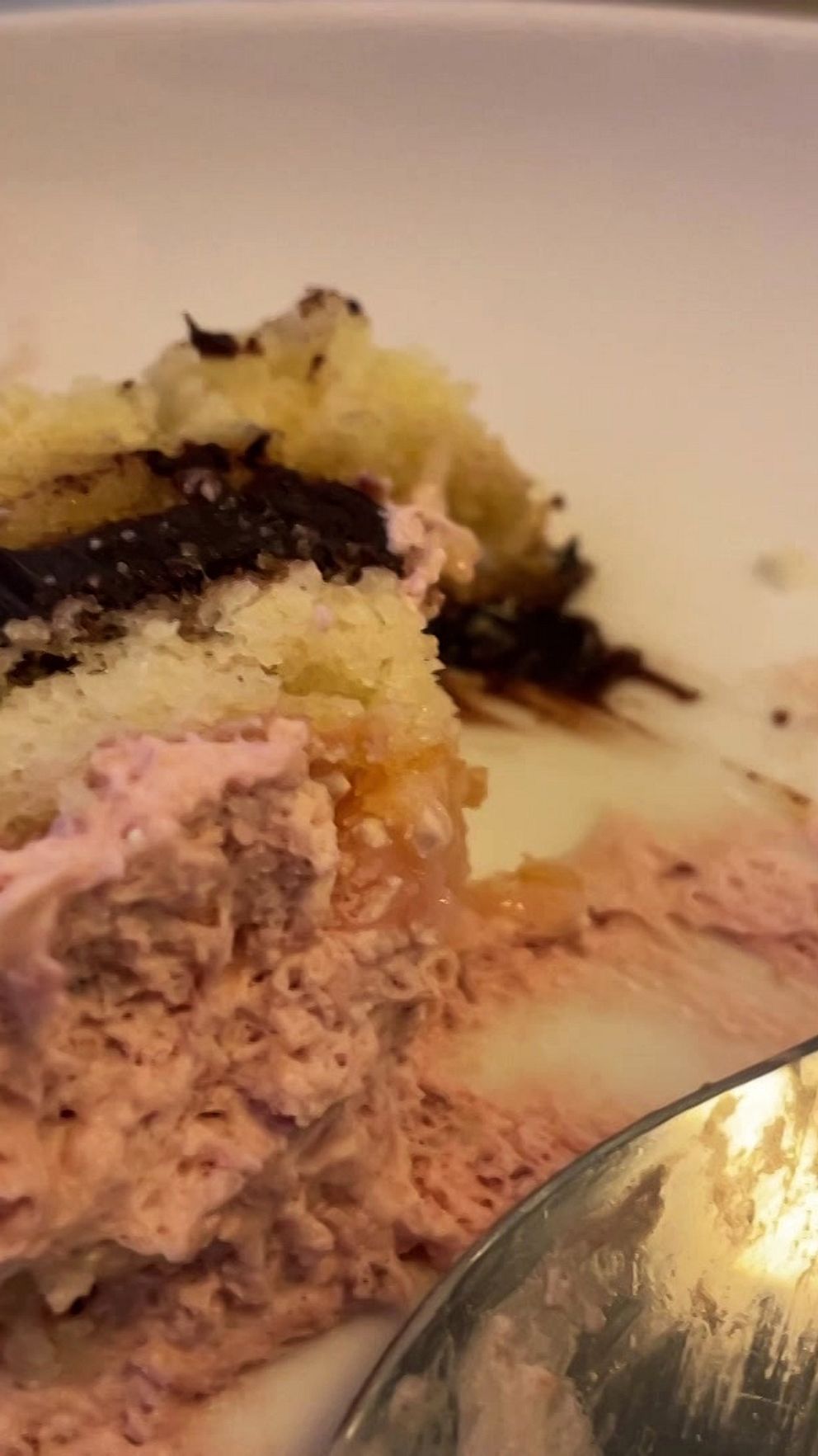 En halväten tårtbit med rosa grädde, chokladöverdrag och ljus tårtbotten på en vit tallrik. En del av en sked syns också i bildens nedre högra hörn.