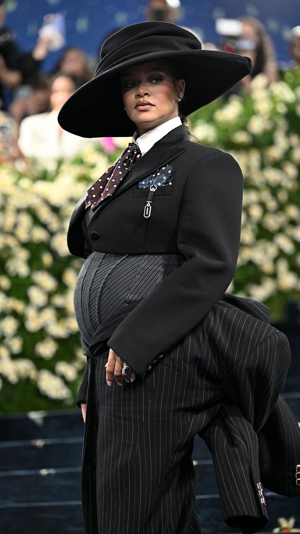 Person i svart kostym med randig väst och stor hatt på en gala, blommor i bakgrunden.