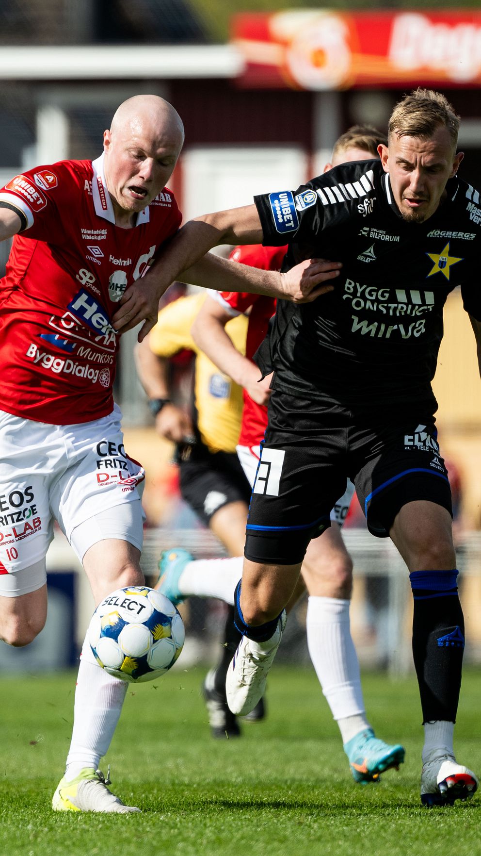 Två fotbollsspelare tävlar om bollen under en match inför publik på arenan.