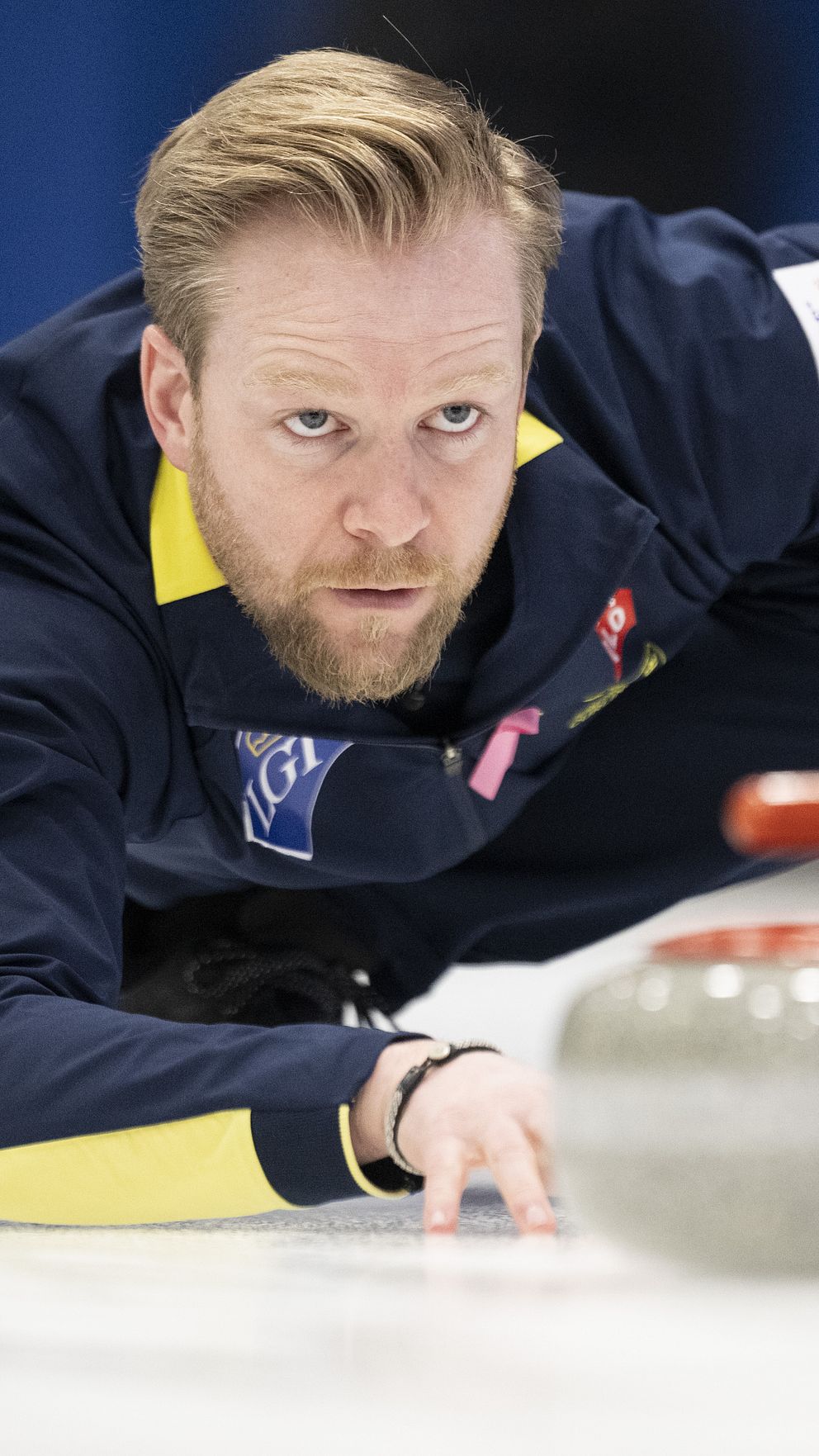 En curlingspelare ligger på knä på isen, sträcker sig fram mot en curlingsten med en snabb rörelse. Denna spelare bär en mörk jacka med gula detaljer och visar en koncentrerad hållning, med fokus på spelet. Curlingstenen ligger nära spelaren, på väg mot målet. Bakgrunden är suddig, vilket framhäver spelaren och stenen.