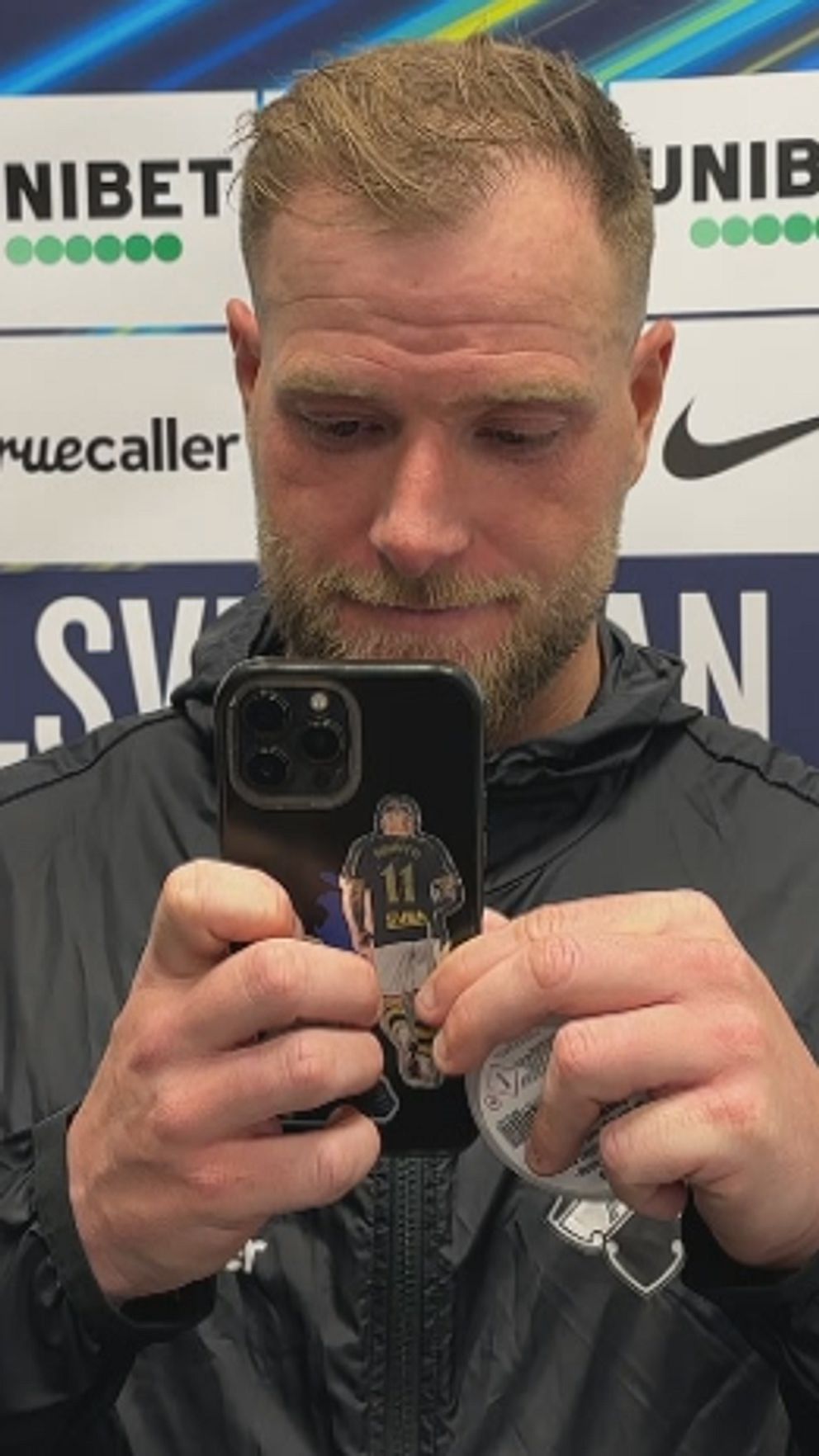 En person i sportjacka håller fram en mobil framför Allsvenskan-vägg.