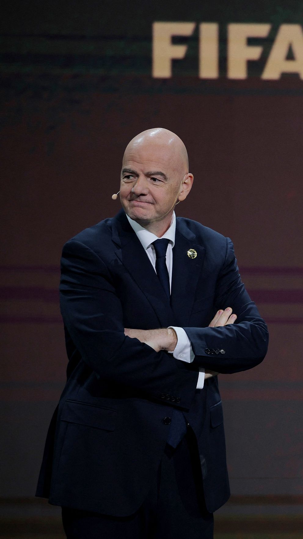 Gianni Infantino, ordförande i FIFA, iklädd en mörk kostym står med armarna i kors framför en bakgrund där ordet ”FIFA” är synligt. Han bär en slips och ett märke på kavajslaget.