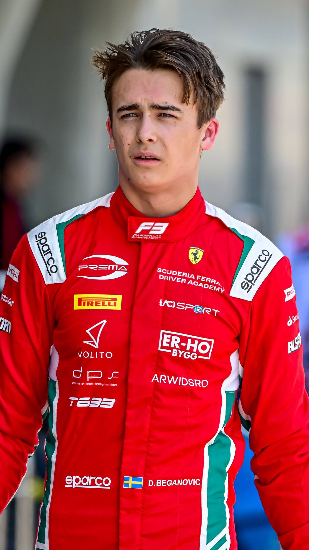 Racerförare i röd Ferrari-dräkt vid racingbana, omgiven av teammedlemmar i bakgrunden.