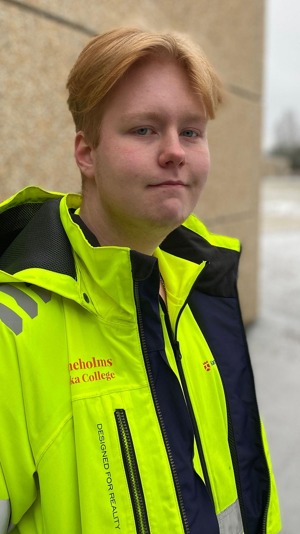 En person står utomhus bredvid en tegelvägg, iklädd en reflexjacka i neongult, vilket indikerar arbete i en arbetsmiljö. Det är vinter med snö på marken och träd skymtas i bakgrunden.