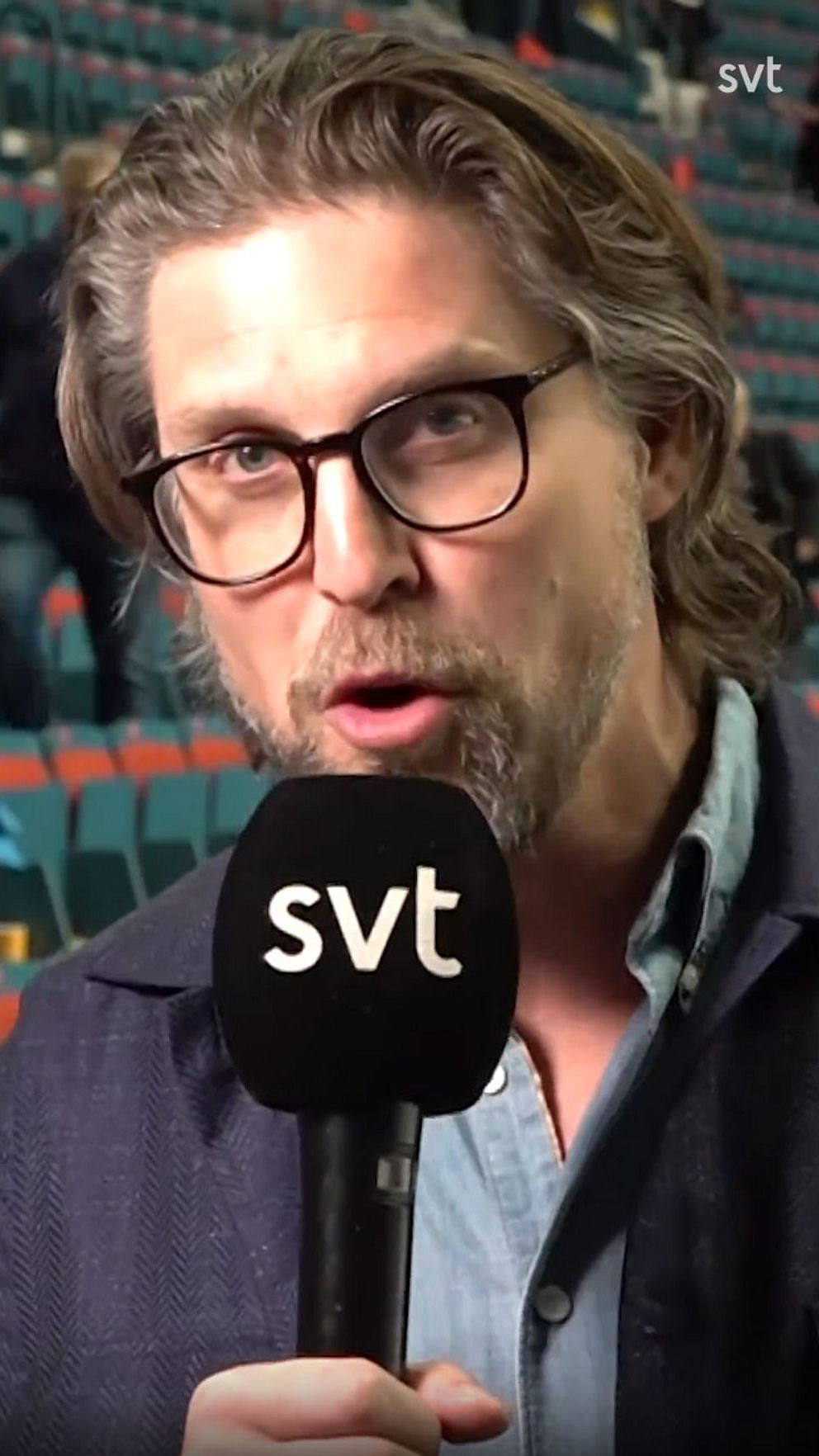 Person håller SVT-mikrofon. Bakgrund: tomma stolsrader i en lokal.