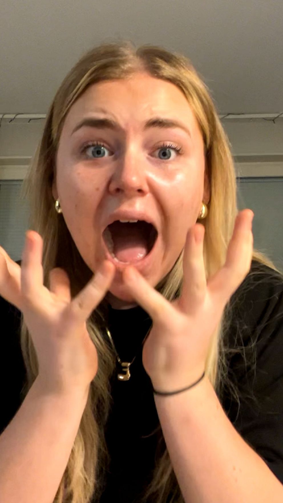 En person med långt, ljust hår och iklädd en svart tröja sitter inomhus framför ett fönster. Personens händer är lyfta, upp och öppna med handflatorna uppåt. I bakgrunden syns ett fönster med gardiner och några ljusslingor.