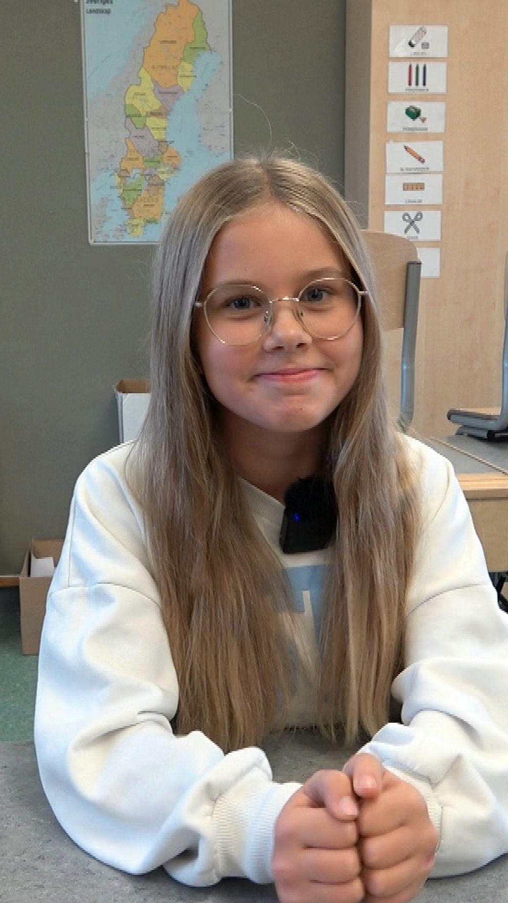 En elev sitter vid ett bord i ett klassrum, med händerna ihoplagda. I bakgrunden syns stolar och bord, samt en vägg med kartor och informationstavlor. Klassrummet har en enkel och disciplinerad miljö, vilket antyder en lärande atmosfär.