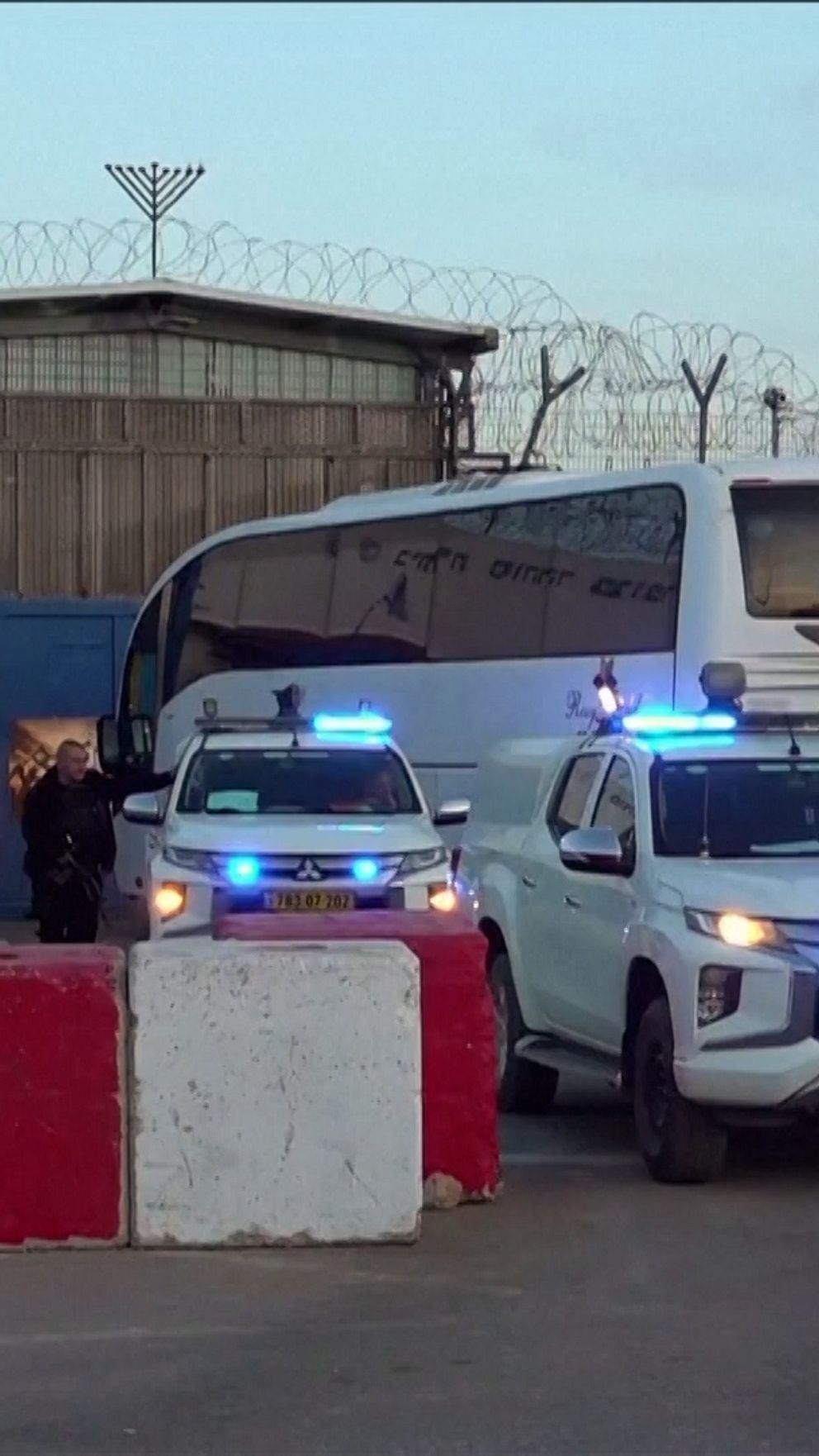 En säkerhetskontroll vid ett fängelse, där en buss och flera polisfordon med blåljus står parkerade. Personer i uniform med vapen övervakar området och ett staket med taggtråd syns i bakgrunden.