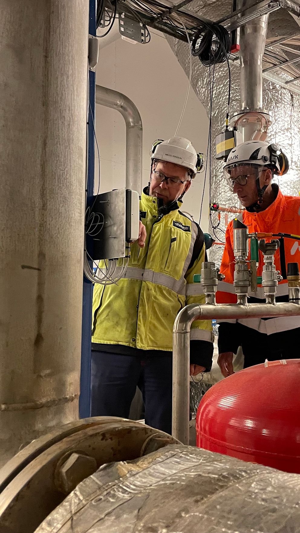 Två personer arbetar tillsammans i en industriell miljö, omgivna av rörledningar och utrustning. Den ena personen bär en gul och svart jacka med hjälm, medan den andra har en orange jacka och hjälm. I förgrunden syns stora metallrör och en röd tank. Bakgrunden består av en isolerad vägg och tak med synliga ledningar.