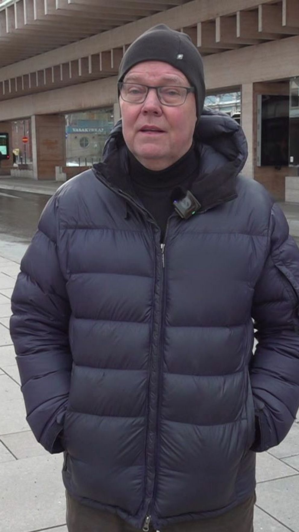 En person i en mörk blustert jacka står på en tom gata med butiker och en modern byggnad i bakgrunden. Gatan är halvmörk, och det finns vinterljus längs trottoaren.