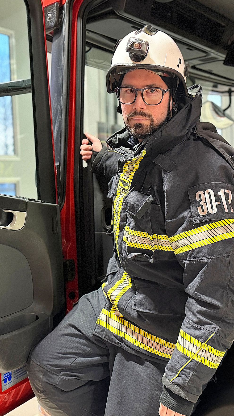 Brandmannen Jon Fredriksson står i dörröppningen till en brandbil, iförd en svart skyddsoverall med reflektorer och en hjälm. Brandbilen står på brandstationen i Mora där räddningstjänsten Norra Dalarna utgår ifrån.