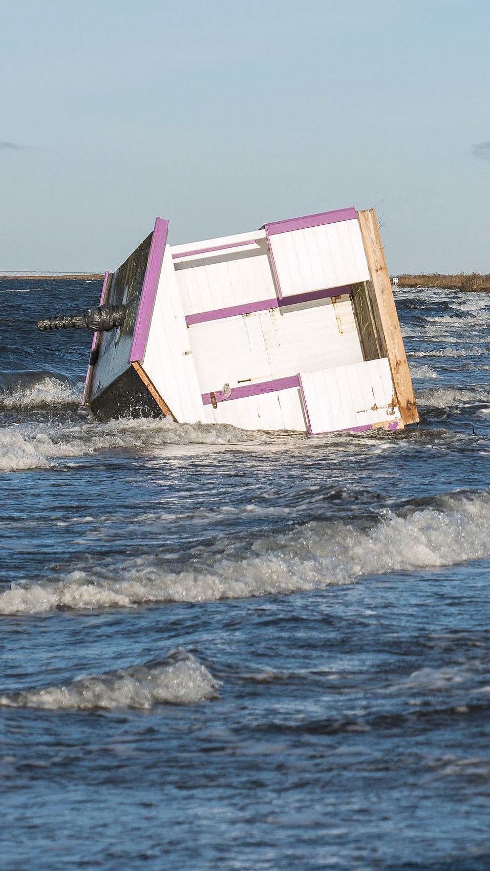 En badhytt har vält och ligger delvis nedsänkt i havsvågor nära stranden.