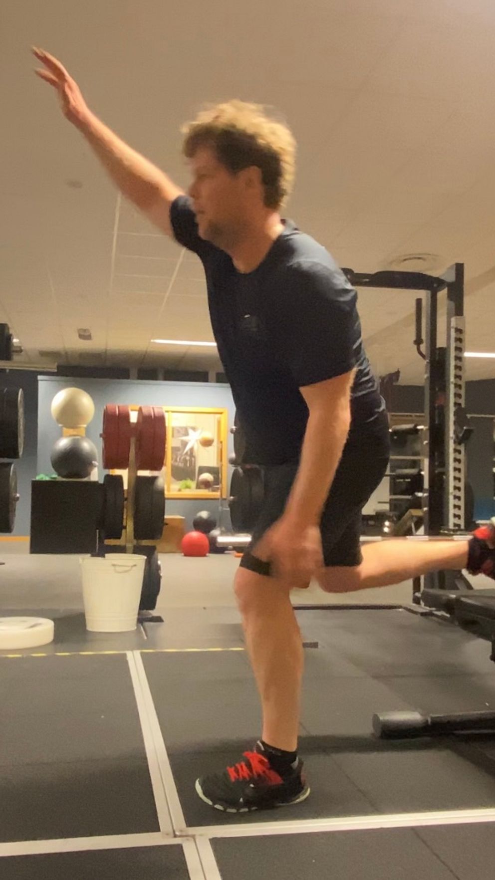 En person utför en balanserad övning i ett gym, med en arm lyft uppåt och den andra benet sträckt bakåt, vilket visar fokus på stabilitet och styrka. I bakgrunden syns gymutrustning och vikter.