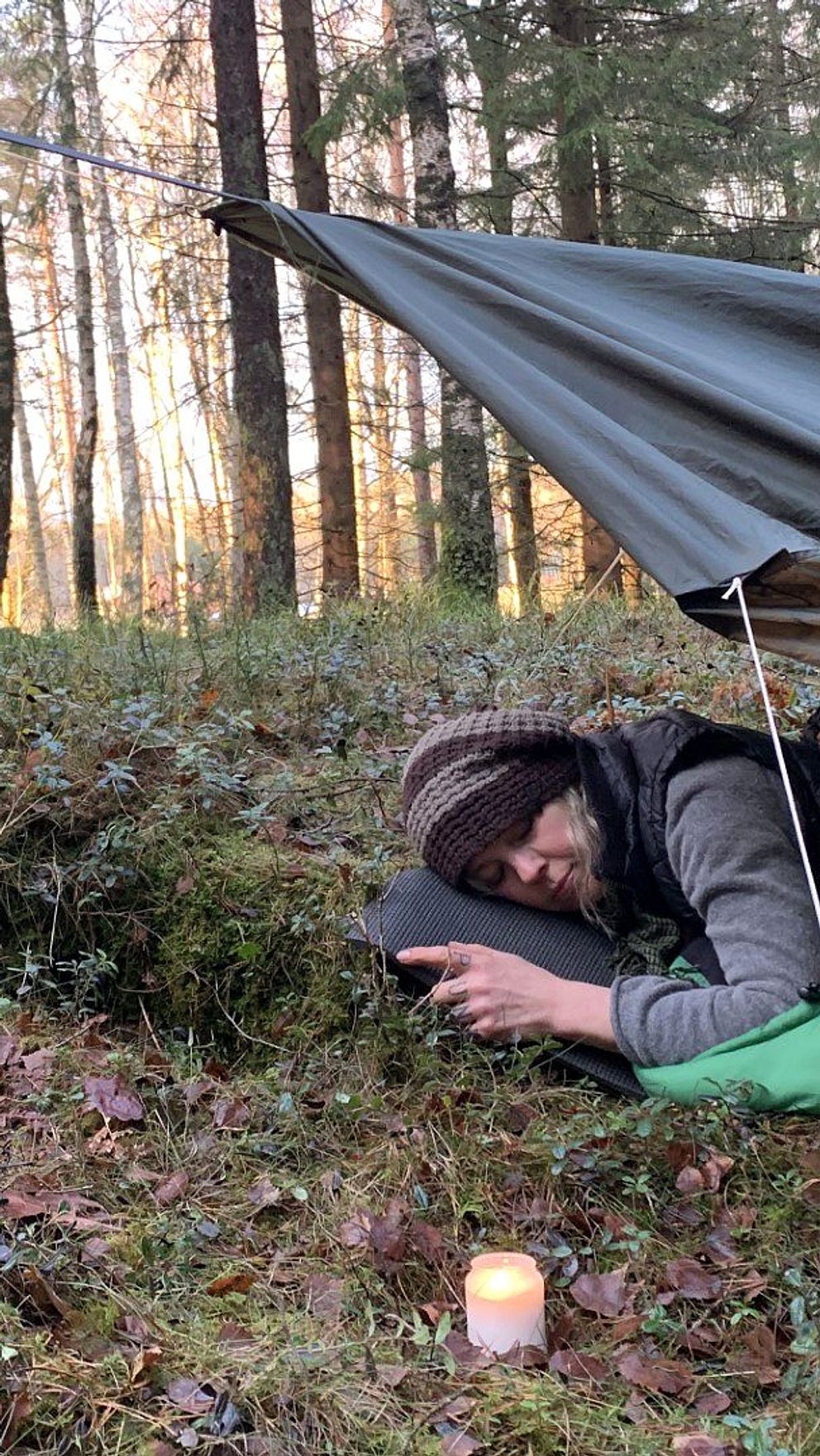 Karin ligger och sover i en grön sovsäck i skogen med ett vindskydd över sig. Bredvid sig har hon ett levande ljus.