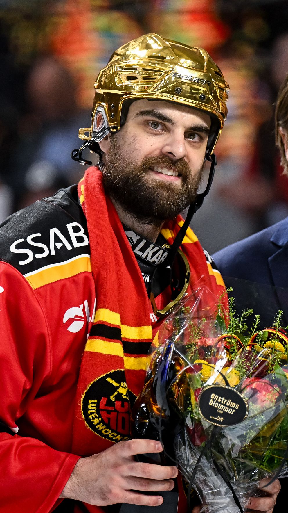 Ishockeyspelare med guldhjälm tar emot blombukett efter match, står bredvid en man i kostym.
