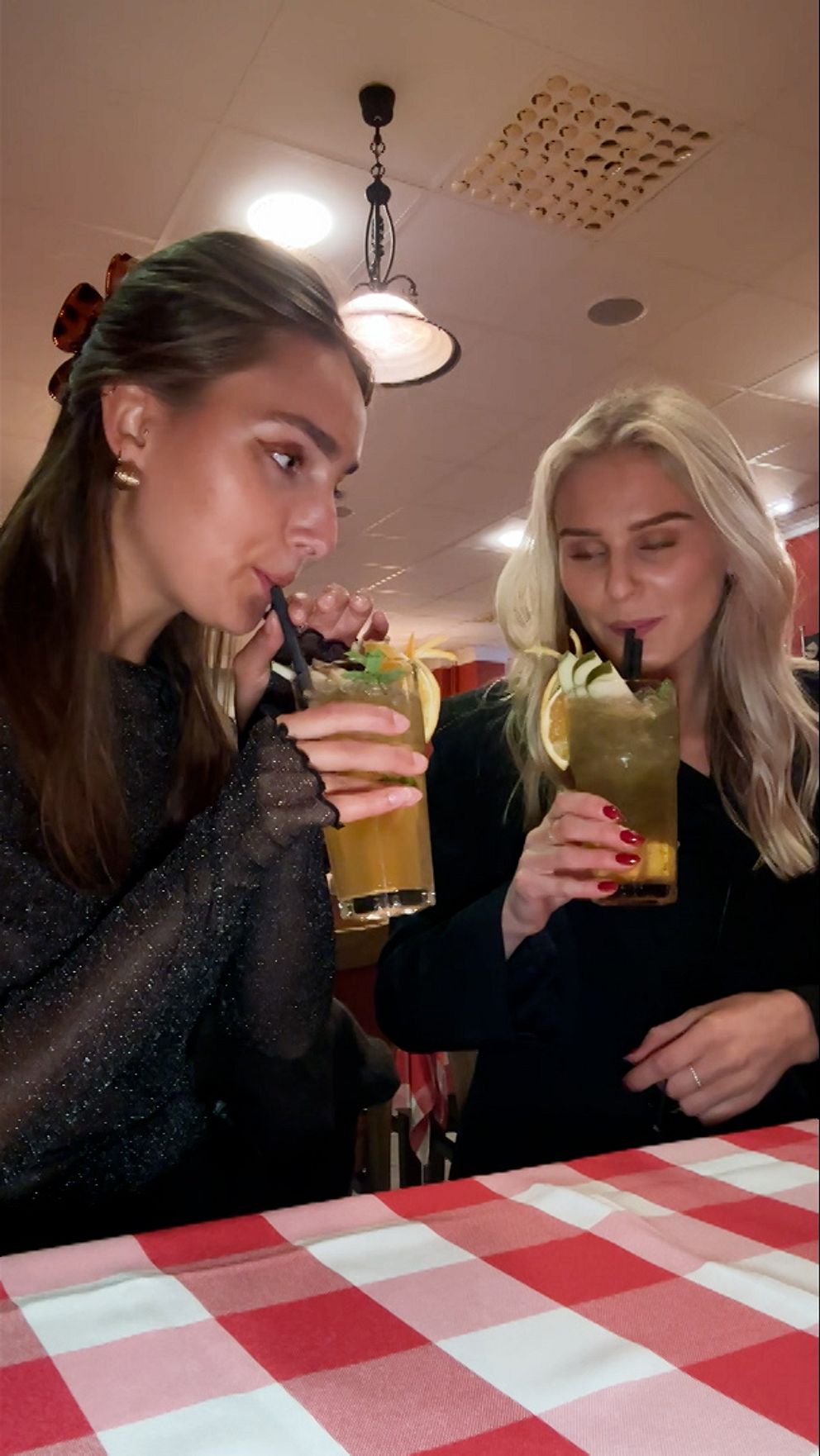 Två personer sitter vid ett bord med en rutig duk. De håller upp glas med drycker och ser glada ut. Bakgrunden visar en inomhusmiljö som verkar vara en restaurang.