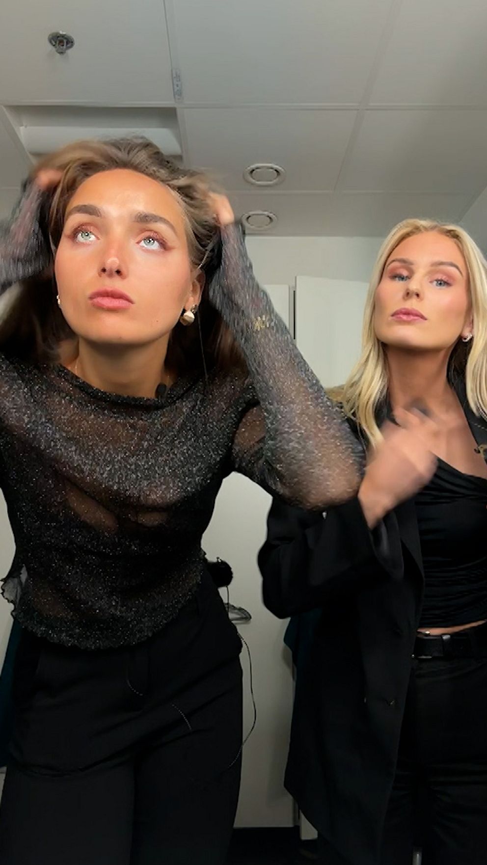 Två personer står i en spegel och justerar sina frisyrer. Den vänstra personen bär en mörk, genomskinlig tröja och svarta byxor, medan den högra personen har på sig en svart outfit.